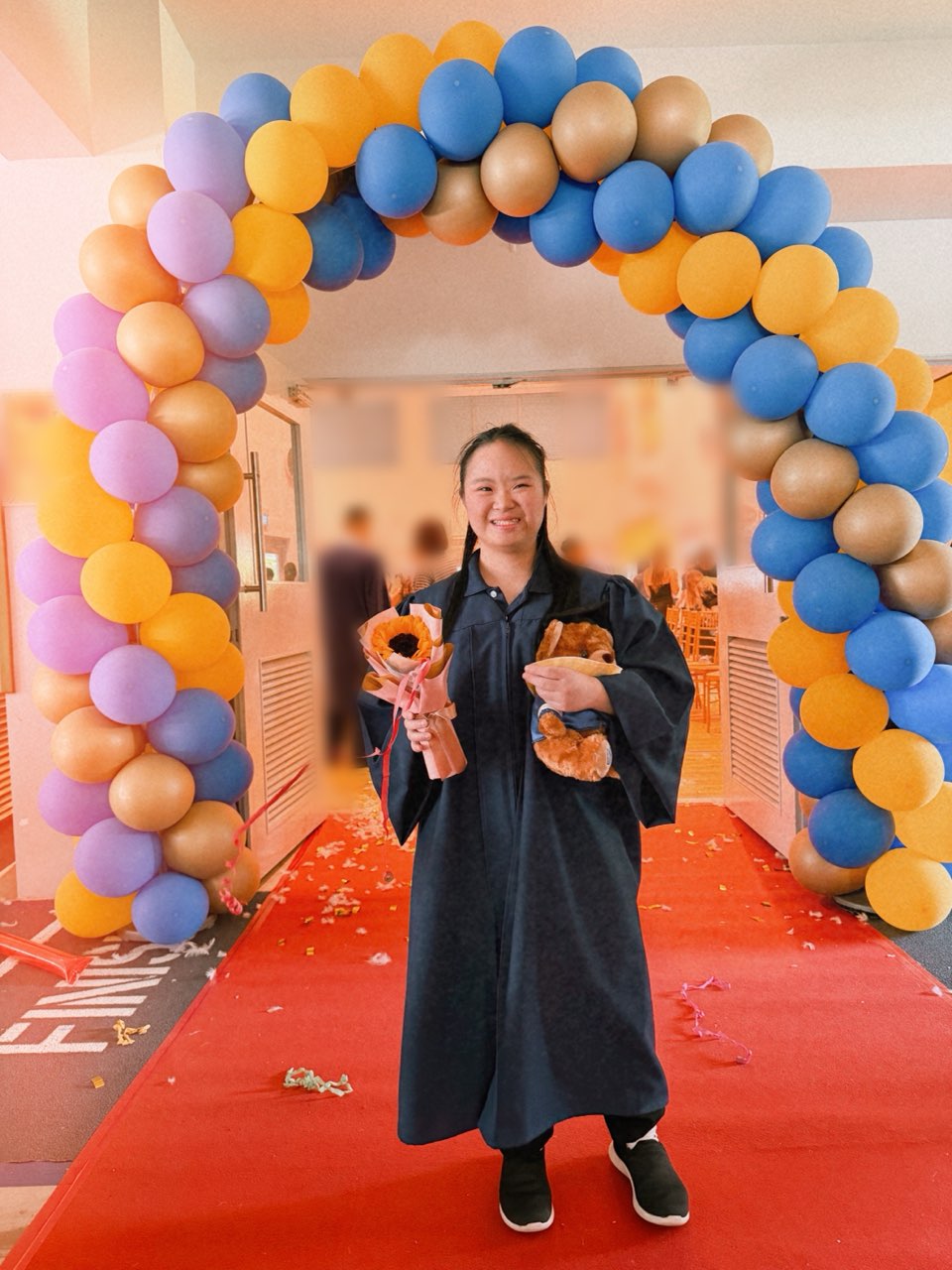 Megan Tang in her graduation gown under a balloon arch, holding flowers and a teddy bear
